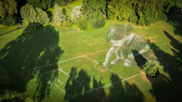 Aerial view of a giant girl drawing on the green field, bathed in soft sunlight, creating a whimsical scene against the backdrop of lush trees, Geneve, Geneve, Switzerland.
