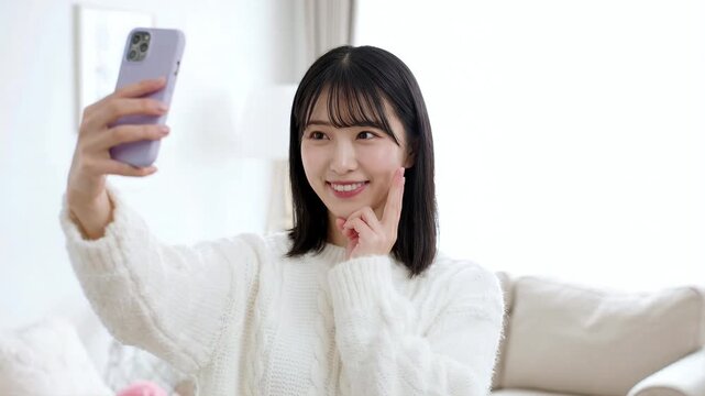 部屋でスマートフォンを使ってピースサインをしながら自撮りをする笑顔の若い女性