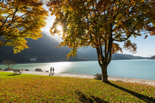 Travel, tourism: Couple walking on the beach of Molveno lake in autumn, enjoying the landscape with colorful trees and mountains at Molveno lake, Trentino, Italy.
