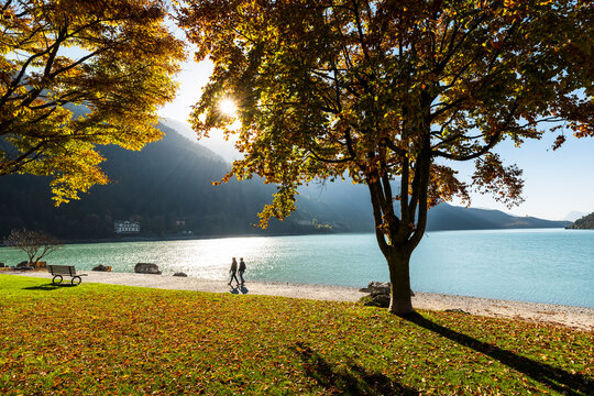 Travel lifestyle: Couple walking on the beach by Molveno lake in autumn, enjoying nature and sunlight, travel destination concept. Molveno lake, Trentino, Italy.