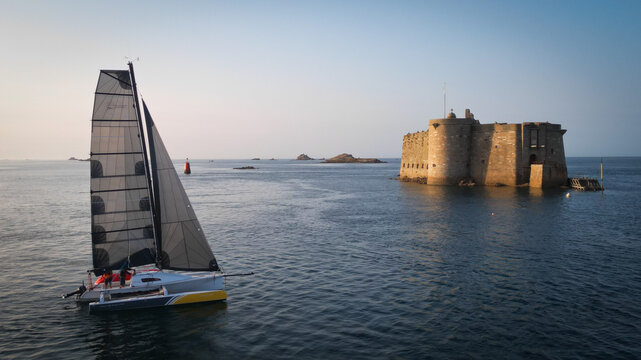 Aerial view of a sleek sailboat gliding past the imposing Chateau du Taureau fortress, its stone walls reflecting the calm sea, Plouezoc'h, Bretagne, France.