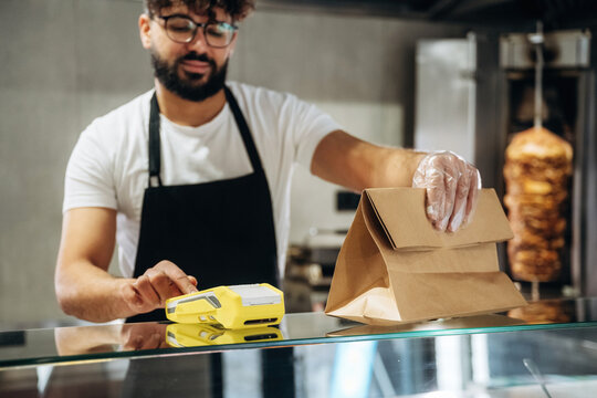 Male food service worker in black apron prepares takeout order with brown paper bag and yellow label printer in a modern restaurant setting