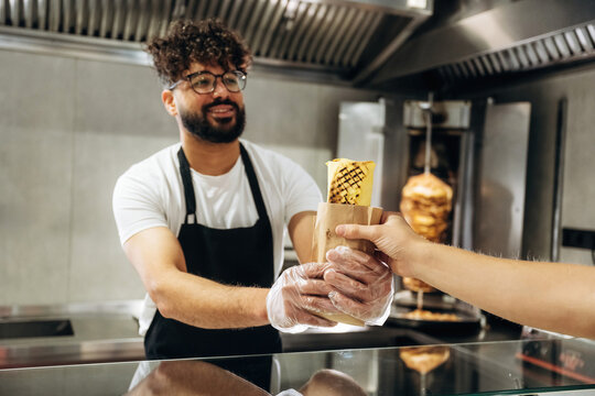 Male food vendor in black apron hands over wrapped kebab to customer at counter in modern fast food restaurant with grill in the background