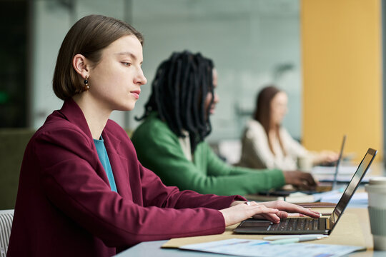 Young businesswoman working on laptop in shared office, collaborating with diverse colleagues. Useful for teamwork, corporate training, productivity campaigns
