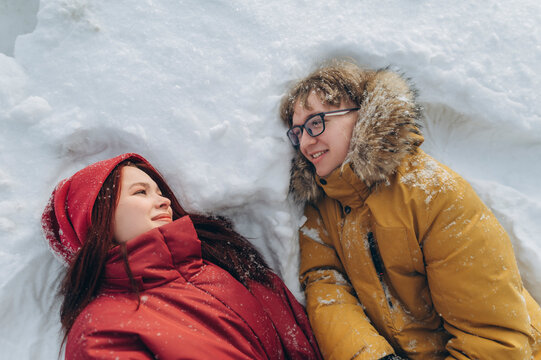 Couple in winter clothing lying in snow smiling at each other outdoors