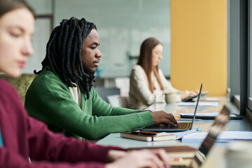 Fototapeta premium Young African American man working on laptop in shared office with colleagues, supporting teamwork and productivity. Ideal for business training, coworking, recruitment, or workplace communication