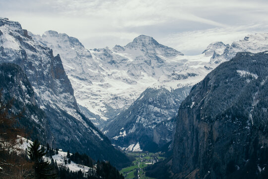 View of snow-dusted peaks towering over a lush valley, where tiny buildings nestle amidst vibrant green fields in the Swiss Alps, Wengen, Switzerland.
