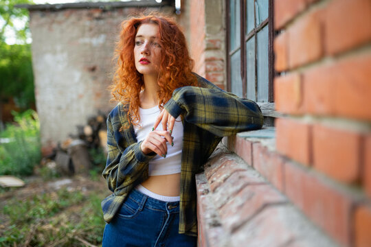 Red haired woman in casual outfit leaning on brick house window