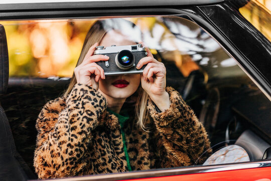 Stylish woman in leopard fur coat taking photo with vintage camera in retro car