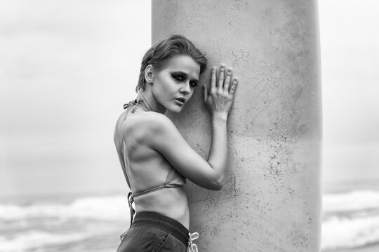 Powerful black and white portrait of woman with pixie cut and intense smoky eye makeup. She leans against textured surface with ocean behind her. The mood is dramatic and moody