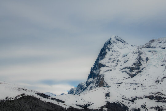 View of a majestic snow-covered mountain peak piercing the cloudy sky, its rugged texture contrasting with the soft clouds, in a serene alpine landscape, Murren, Switzerland.