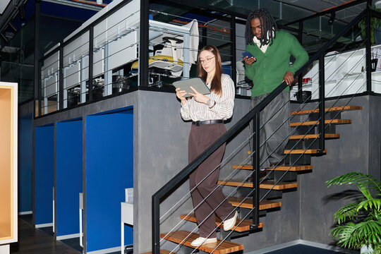 Wide shot of young businesswoman and African American businessman descending office stairs while using tablet and smartphone. Ideal for hybrid work, team communication, productivity