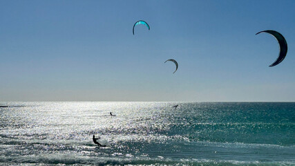 Silhouette of a kitesurfer riding the waves in the turquoise ocean under a clear blue sky, with three colorful sails in the background. Freedom and adrenaline. © Sonja