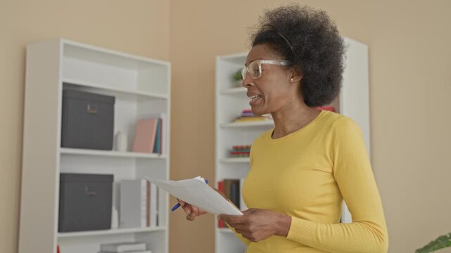 Woman wearing yellow sweater and clear glasses holding papers and gesturing with pen in building; teaching focus instruction.