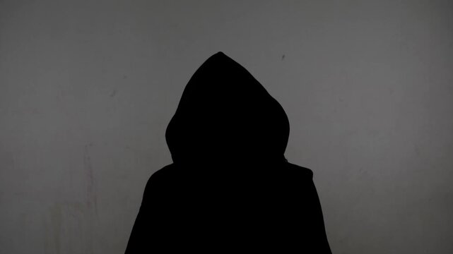 An anonymous silhouette of a person in a hoodie gives an interview against a white, dirty wall