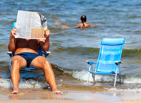 A man is relaxing in an armchair right by the waves, engrossed in his reading and covering his face with an open newspaper