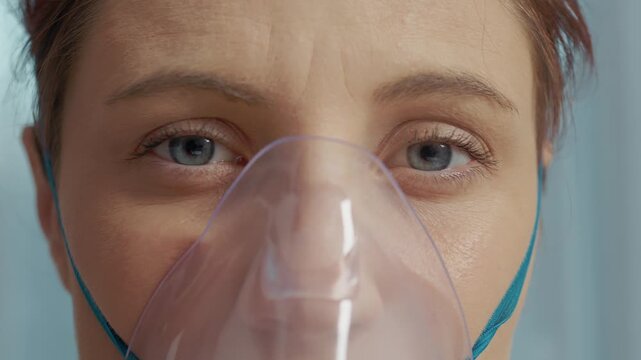 A woman sits at home wearing a mask connected to a nebulizer. She inhales steam from saline to help with cough and respiratory issues while her eyes are focused.