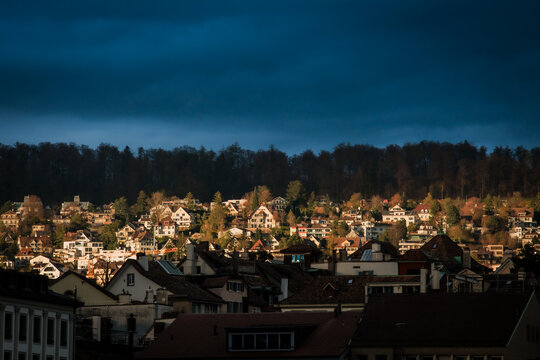 Zurich, Switzerland - 02 April 2023: View of buildings scattered across the landscape, a tapestry of light and shadow playing upon their rooftops, set against a backdrop of dark, dense forest.