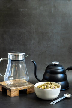 Yerba Mate loose tea in a white ceramic bowl with kettle and tea pitcher on a dark backdrop