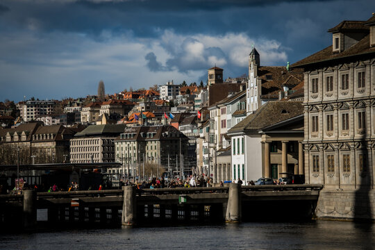 Zurich, Switzerland - 02 April 2023: View of the Limmat River flowing under the Quaibrucke bridge, with the historic architecture of the city rising towards a dramatic sky.
