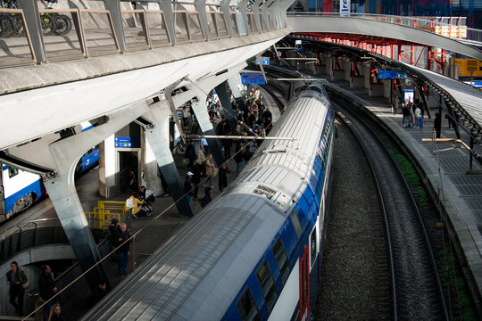Zurich, Switzerland - 02 April 2023: View of a bustling train station where a sleek, silver train glides along the tracks beneath the modern, angular architecture and throngs of people.