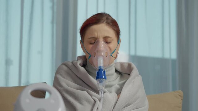 A woman is using a nebulizer with a mask to inhale a saline solution for her cough. She is sitting indoors while treating her respiratory infection.