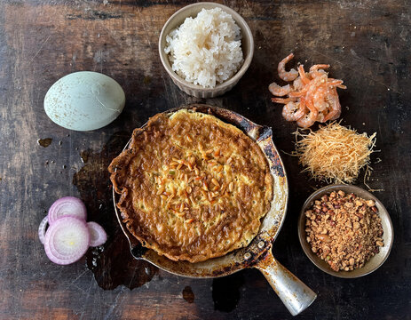 Overhead View of Kerak Telor Betawi Cooked in Rustic Iron Pan, Surrounded by Duck Egg, Glutinous Rice, Dried Shrimp, Shredded Coconut, Peanuts, and Sliced Shallots on Weathered Wooden Surface