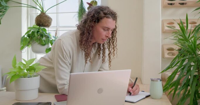 Nonbinary person leaning forward, writing in notebook while working on laptop and phone at counter