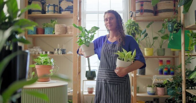 Adult African nonbinary in apron at plant shop holding two pots as camera focusing, showing plants