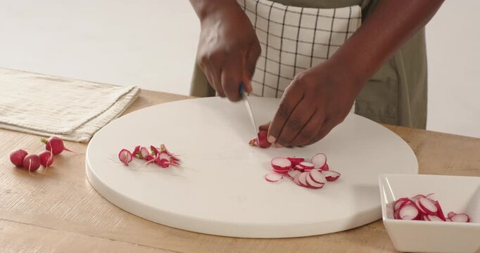 Chef's knife is slicing thin radish rounds on white cutting board on wooden table, filling bowl
