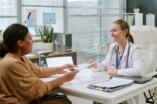 Medium shot of Caucasian female doctor handing medical document to Black woman patient during consultation in clinic