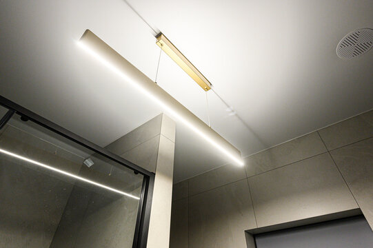 A modern bathroom with a suspended linear ceiling light, neutral-toned tiles, a glass shower enclosure with LED strip lighting, and a ceiling vent. Minimalist design, clean lines.