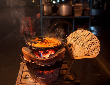 Traditional Clay Anglo Brazier with Kerak Telor Cooking Over Glowing Red Charcoal at Night, Betawi Street Food Vendor Scene with Woven Bamboo Fan and Dramatic Fire Light