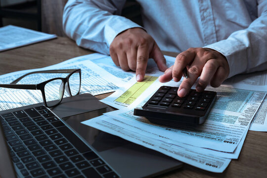 Business professional calculating financial data with calculator and laptop, analyzing documents, budgeting expenses, tax planning, accounting and finance concept in modern office workspace.