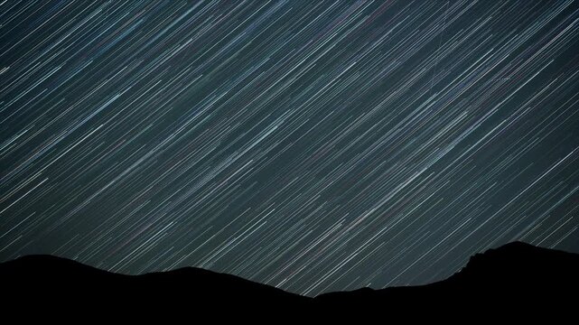 Multicolored star trails arcing above a distant mountain landscape in a stunning night sky time lapse of cosmic motion.