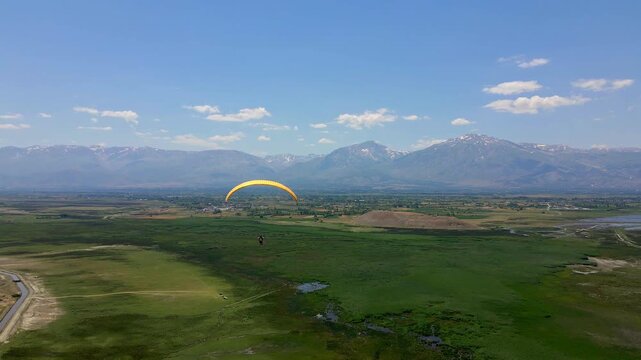 Paraglider soars above lush green fields. Mountains loom distant under clear blue sky. Calm flight evokes freedom and adventure. Perfect for travel, nature, or outdoor lifestyle themes