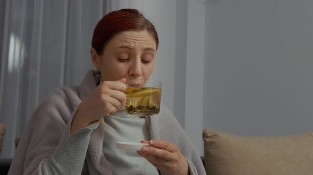 A woman sits on her couch wrapped in a blanket. She holds a cup of herbal tea and uses tissues for her cold symptoms. She is trying to feel better and rest.