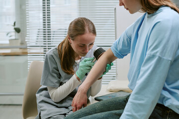 Young adult Caucasian female clinician examining woman arm with handheld diagnostic device during dermatology checkup © AnnaStills