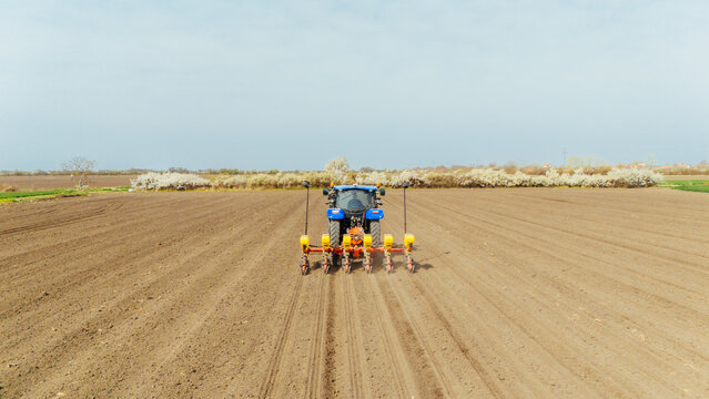 Aerial view of a blue tractor planting seeds in the brown earth, with a backdrop of blooming trees under the muted sky, Sremska Mitrovica, Vojvodina, Serbia.