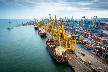 Obraz premium Containers are loaded and unloaded at the cargo sea port. Cranes move goods while large ships dock along the waterfront. Workers manage operations on the ships and wharf