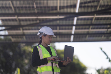 Fototapeta premium A female technician wearing a safety helmet and reflective vest uses a laptop while inspecting a solar carport installation. The scene features solar panels and parked cars under a clear sky.