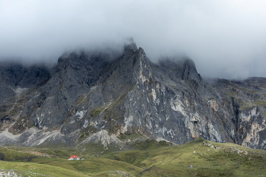 View of a solitary red-roofed house nestled in the vibrant green foothills beneath towering, jagged mountains shrouded in ethereal mist, Picos de Europa, Asturias, Spain.