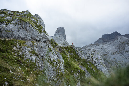 View of a solitary figure stands atop a rugged, grassy ridge amidst the imposing, gray peaks of a mountain range under a cloudy sky, Picos de Europa, Asturias, Spain.