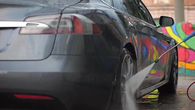 Male hand washing a modern electric auto at a self-service car wash using a high pressure water stream. View captures dynamic splashes highlighting eco friendly transport and urban routine. Slow mo