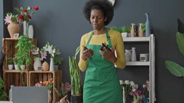 Woman in green apron holds pruning shears in hands and inspects stems and vases among shelves of flowers in a floral studio; quiet concentration.