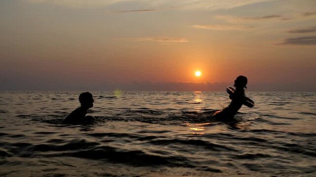 Teenage boy and girl play in sea at sunset throwing ball and making splashes. Brother and sister happy play in water on ocean coast. Happiness to be together and love each other Golden sunset on coast