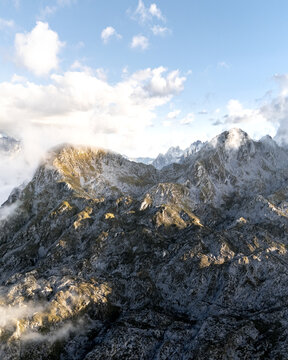 Aerial view of rugged, rocky peaks pierce through a veil of ethereal clouds under a bright sky, Picos de Europa, Asturias, Spain.