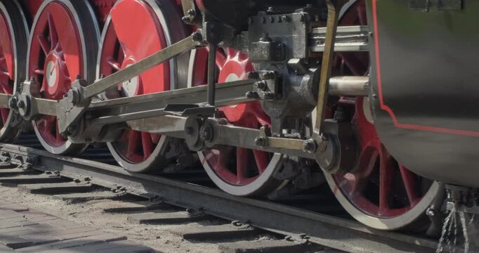 A genuine vintage steam locomotive passes by the camera along the railroad tracks. The levers of the drive mechanism are working, the wheels are turning, and the rails, sleepers, and steam are visible