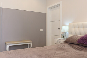Naklejka premium A minimalist bedroom with a beige quilted bed, white tufted headboard, nightstand with a pleated lamp, a light beige bench, and a neutral-toned wall. Soft, calming palette.