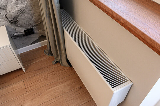 A white wall-mounted convector heater beneath a wooden window sill, adjacent to a beige wall, with wooden flooring, a grey curtain and a white bedside cabinet nearby.